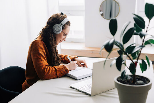 Busy young woman university student wearing headphones using laptop e-learning, writing notes, studying online education seminar class by webinar, learning online seminar sitting at the home table.