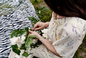 Woman making head wreath with wild flowers. Close up of hands weaving a wreath with green leaves...