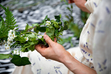Woman making head wreath with fresh flowers. Close up of hands weaving a wreath with green leaves...