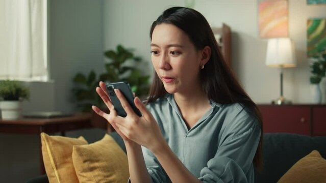 Happy beautiful asian woman sit on couch holding mobile phone in hands chatting and playing social media. Attractive girl looking at smartphone, cellphone, browsing internet on sofa home living room