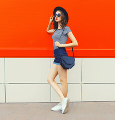 Beautiful woman model posing wearing black round hat, shorts and handbag in the city on orange...