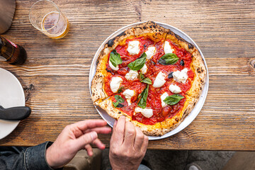 Male hands cutting a freshly baked Neapolitan Margherita Pizza on a rustic wooden table in a traditional Pizzeria.