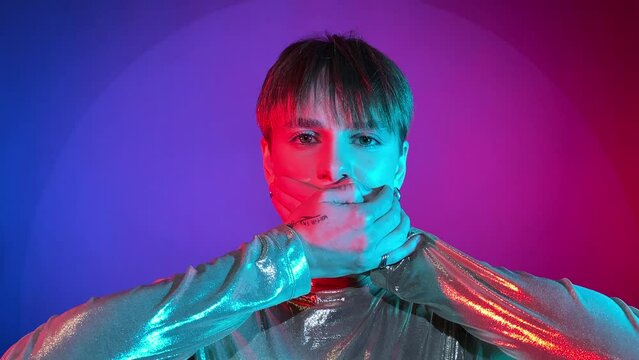 Portrait young woman covering mouth with hands, showing speak no evil gesture, looking camera neon illumination indoors. Studio shot art colorful lighting. Female short hair wearing sparkling clothing