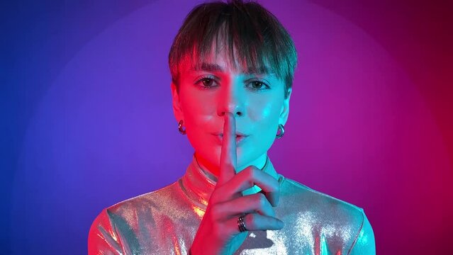 Portrait young adult woman saying hush be quiet with finger on lips shh gesture in neon illumination indoors. Studio shot in art colorful lighting. Female person short hair wearing sparkling clothing