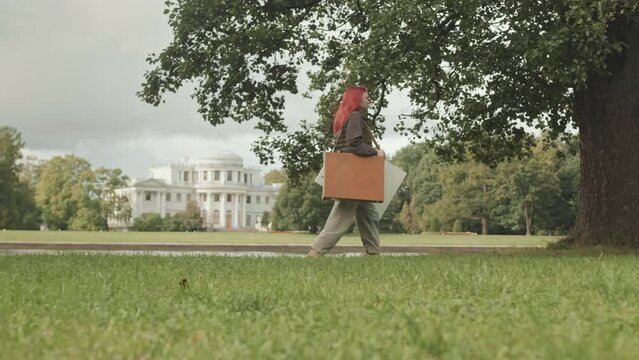 Full length side view shot of red haired female artist with canvas and paints in wooden case walking in park looking for place to paint