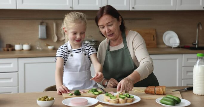 Elderly loving granny and cute preschooler girl grandchild in aprons, talking, cook together in kitchen, put meat and cheese on bread slices, prepare sandwiches for family breakfast at home. Cooking