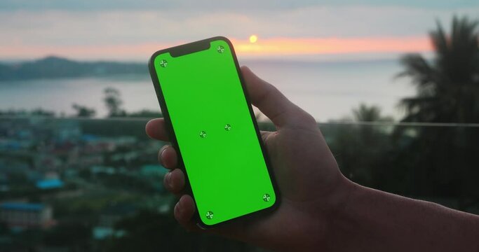 Close up male holding phone with green screen chroma key over sunset at sea at tropical scenery. Male hand with phone with mobile green screen mockup on tropical beach at sunset on summer vacations