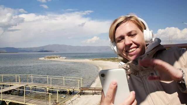 A pretty adult young woman is using a smartphone for a video call and talking on the shore of a beautiful mountain lake while on a trip. Videoconferencing using a mobile phone while on vacation
