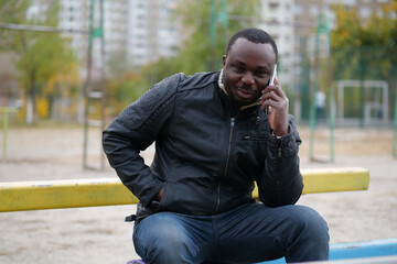 African american man sits at sport ground and speaks on smartphone