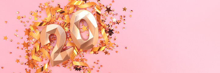 Banner with gold colored number 20, ribbons and stars confetti on a pink background.