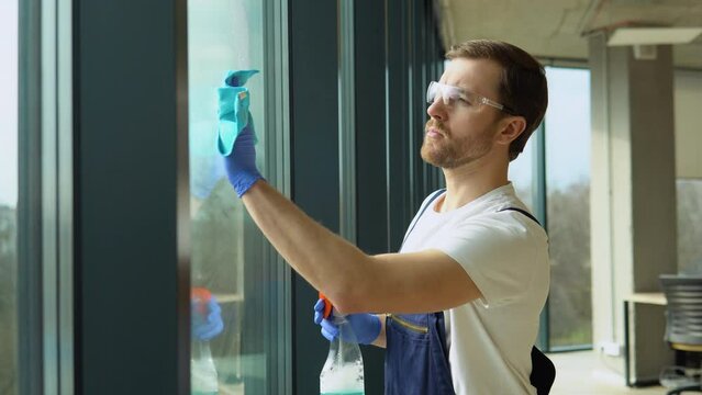 Male cleaning service worker in uniform and protective glasses washing office windows