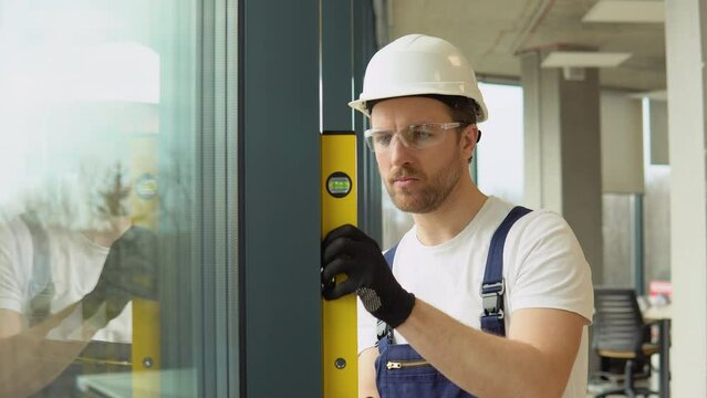 A repairman in uniform installs pvc windows in new office. A man builder measures of the vertical deviation of pvc windows