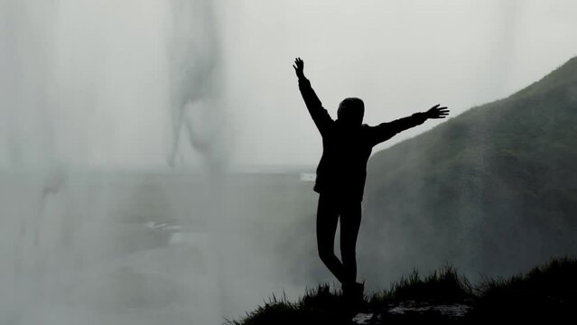 Woman standing on the edge of the cliff and raising her hands up in the air wiht beautiful Detifoss waterfall in background. Iceland