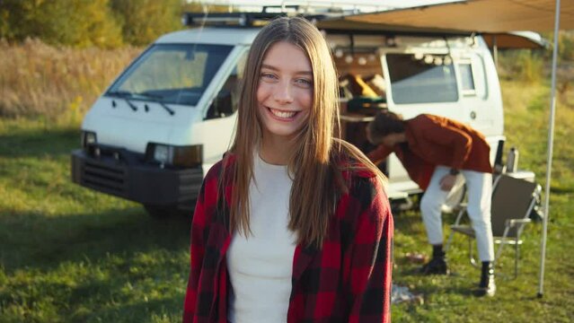 Beautiful picture of attractive young caucasian girl looking away, turning her head, and smiling at the camera. Camping site, white van, young man in background. Picnic in nature, daylight