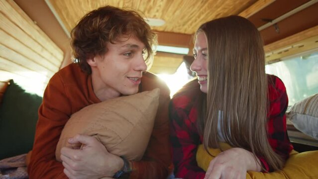 Beautiful caucasian couple lying on a bed in a small cozy camper van and talking. Young hipster girl kisses her boyfriend on the cheek. Romantic road trip, outdoor weekend