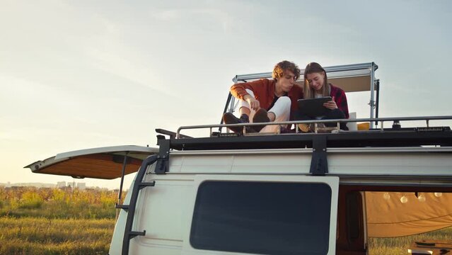 Young caucasian couple sitting on camper van rooftop, snacking and using a tablet on their road trip. Boy and girl resting on a backcountry picnic. Evening, blue sky, countryside background