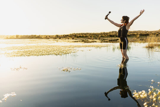 Woman taking selfie with camera in nature in middle of lake, surrounded by flowers, with selective focus, sunset in middle of lake woman traveling alone, waves in water, movie colours.