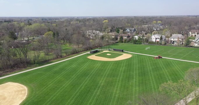 Aerial view of a baseball and soccer field in a large suburban park with a sports field mower tractor grooming the grass in early spring.