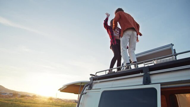 Beautiful caucasian pair dancing on their camper van rooftop. Young girl and boy enjoying their romantic road trip. Countryside background, summer evening, relaxing on fresh air