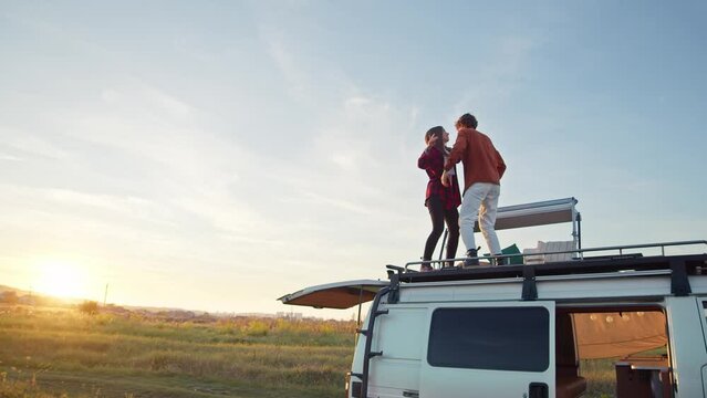 Beautiful caucasian pair dancing on their camper van rooftop. Young girl and boy have fun on a romantic road trip. View from a distance, countryside background summer sunset, rural retreat
