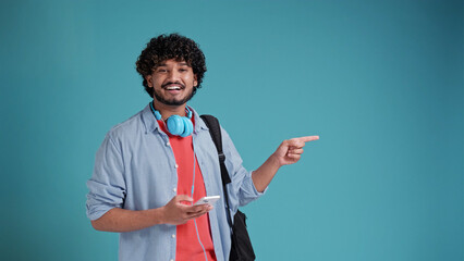 young latin spanish man with headphones and smartphone pointing with his finger to the side to the space for text, on a blue studio background