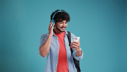 indian or spanish young male student listening to music with headphones and smartphone on blue studio background