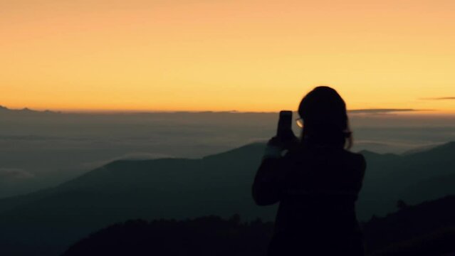 Back view of caucasian woman hand holding phone and taking photo on the red sunset at the camping in the mountains. Evening sky scene with golden light from the setting sun in summer