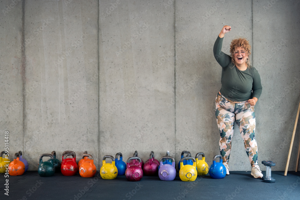 Wall mural Beautiful young smiling plus size woman in fitness clothes feeling confident and happy in her body, satisfied after workout session raising hand in yes gesture rejoicing success in gym. Copy space