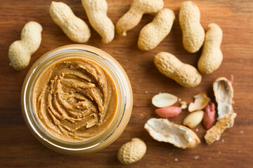 Creamy smooth peanut butter in jar with peanuts in shell around, photographed overhead on wood...