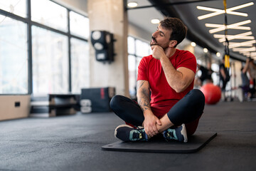 Fototapeta na wymiar Athletic man in sports clothes warming up for sports training in gym, stretching neck muscles before workout, sitting on exercise fitness mat.