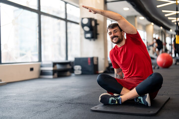 Young motivated sports man doing upper body exercise, stretching his arm and core muscles, sitting on floor on exercise mat, warming up before sports training session in gym.