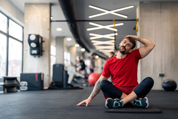 Fototapeta na wymiar Young male athlete doing stretching exercise in gym while sitting with crossed legs on exercise mat. Sportsman in red t-shirt stretching neck muscles during warm up session, sitting on floor in gym.