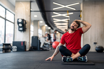 Obraz na płótnie Canvas Athletic man in sports clothes warming up for sports training in gym, stretching neck muscles before workout, sitting on exercise fitness mat, breathing with eyes closed.