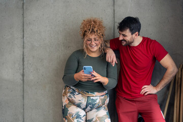 Young confident curvy woman tracking exercise progress in fitness center over smart mobile phone with her fitness instructor.