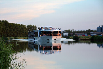 A large tourist passenger ship sails along the calm river between the banks