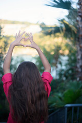 Obraz premium A view of the back of a heart shaped hands of young girl with long hair, pink shirt. Heart made by kids hand at sunset. Tropical view, palms around