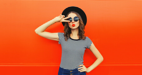 Portrait of beautiful young woman blowing her lips sending sweet air kiss wearing black round hat on orange background