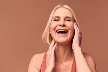 Live photo of a happy older woman laughing and touching her face with her hands while posing in front of the camera. Woman's beauty, facial skin care.Beige background.