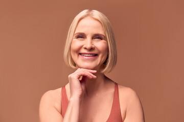 Close-up portrait of a happy beautiful blonde senior woman on a beige background touching her chin...