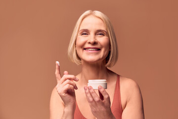 Close-up portrait of a happy beautiful blonde older woman holding a jar of cream in one hand and cream on her finger in the other. Woman laughing and looking at camera on beige background.