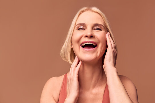 Live Photo Of A Happy Older Woman Laughing And Touching Her Face With Her Hands While Posing In Front Of The Camera. Woman's Beauty, Facial Skin Care.Beige Background.
