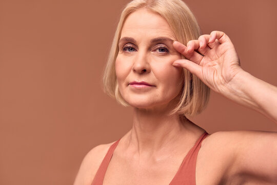 A beautiful blonde older woman on a beige background shows wrinkles near her eyes. Skin aging and skin care concept.
