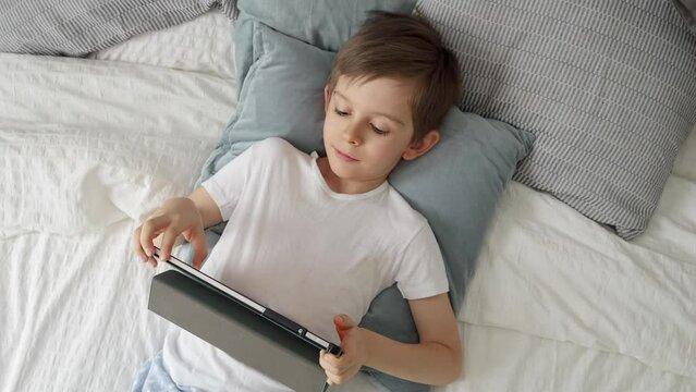 Happy young boy is playing with a tablet computer while lying in bed during the day. Child using gadget, education and development