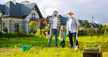 Portrait of happy family at cottage in village. Old father, son and grandson planting tree at...