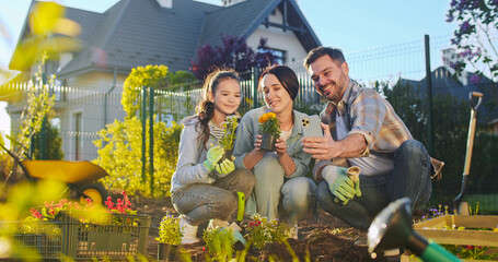 Happy Caucasian family with flowers in pots smiling to camera and videochatting on smartphone. Working in orchard. Father, moher and daughter planting plants and having videochat via mobile phone.