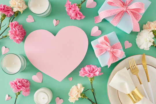 Trendy Mother Day table setting idea. Top view photo of plate with cutlery napkin gift boxes candles carnation flowers hearts on turquoise background. Flat lay with blank heart for text or advert