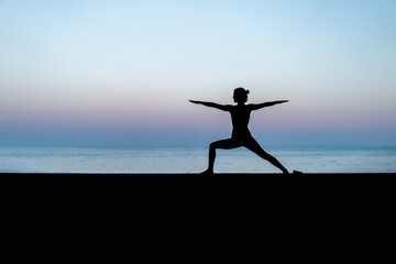 Silhouette de femme faisant du yoga devant la mer au coucher de soleil,  posture Virabhadrasana II...
