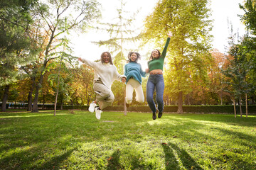 Three young multiracial girls jump happily outdoors in the park. Happiness and joy women with friends on vacation on a sunny spring day. Excited college people celebrating a community success. 