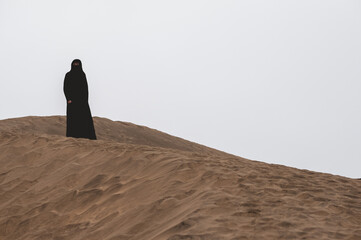 The burqa's mystery: alone muslim woman with black burqa in the vast desert under pale sky