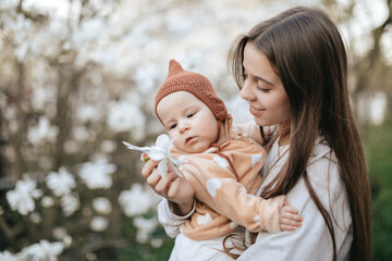 A teenage girl is showing her newborn sister a flower from a magnolia tree in a blooming garden.
